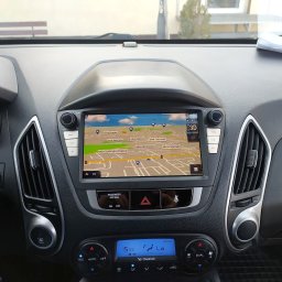 JanSerwis.pl - Wnętrze samochodu z włączoną nawigacją GPS, wyświetlającą mapę ulic. Widoczny panel klimatyzacji z temperaturą 15.5 stopni Celsjusza, przyciski sterowania i kluczyki w stacyjce.