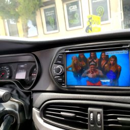 JanSerwis.pl - Wnętrze samochodu z włączonym ekranem radia samochodowego, na którym wyświetlany jest teledysk z pływającymi osobami.