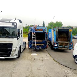 JanSerwis.pl - Serwisant na drabinie przy dwóch otwartych śmieciarkach, biała ciężarówka z przodu, firmowy van z reklamą serwisu obok, szare niebo.