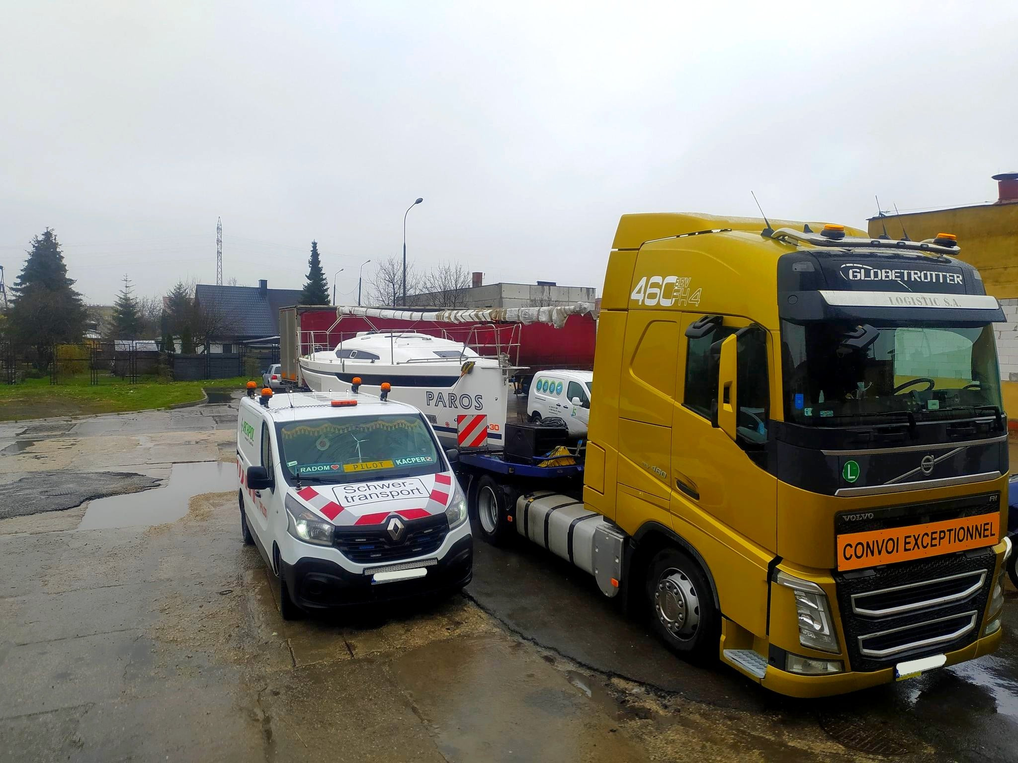 Żółta ciężarówka Volvo z napisem 'CONVOI EXCEPTIONNEL' przewozi łódź żaglową 'PAROS' na platformie, eskortowana przez biały samochód pilotujący 'Schwer-transport' z napisem 'PILOT Kacper' na mokrym...