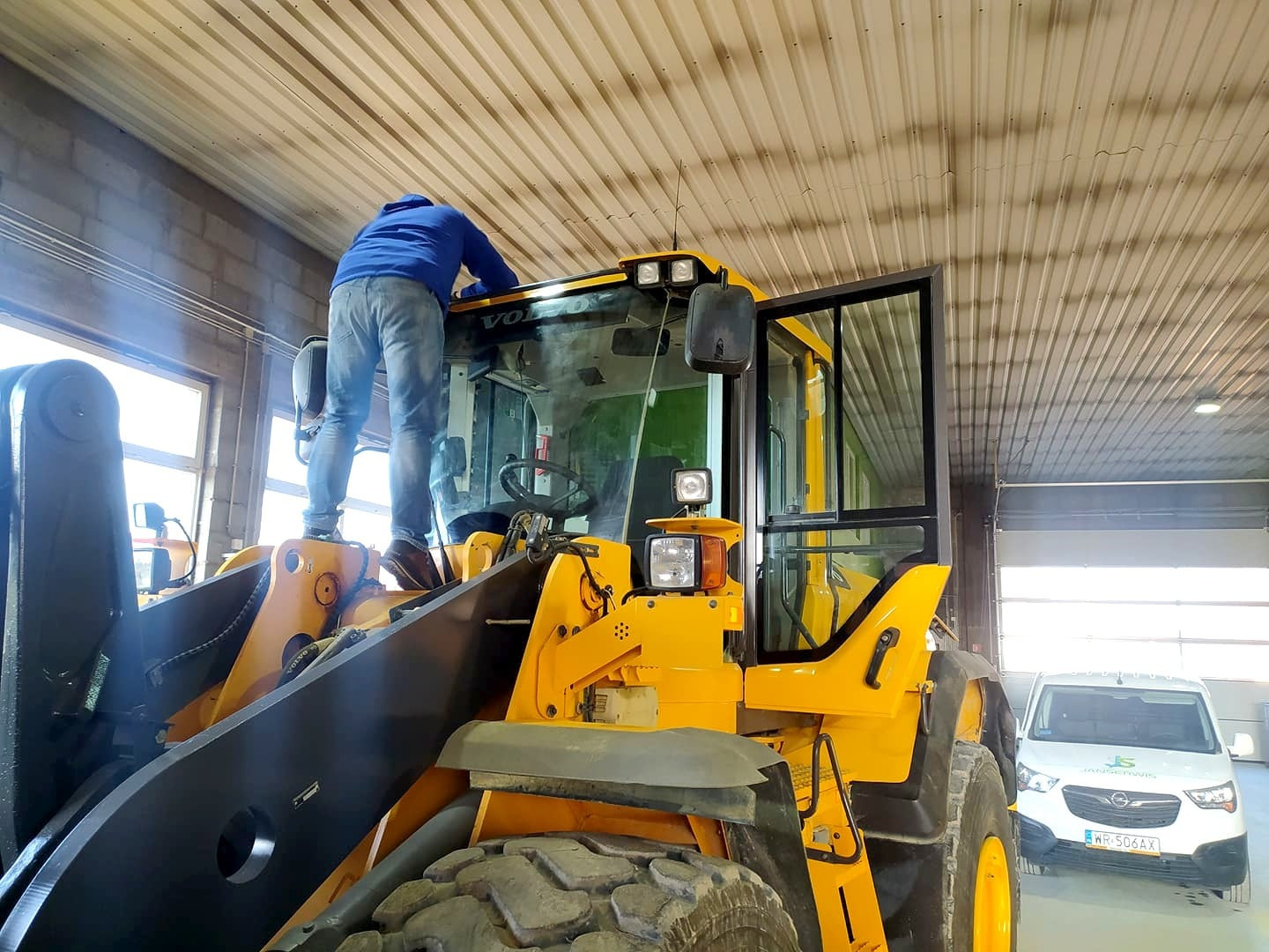 Mężczyzna w niebieskiej bluzie stoi na żółtej ładowarce Volvo w warsztacie, prawdopodobnie podczas naprawy lub konserwacji. W tle widoczny biały samochód dostawczy.