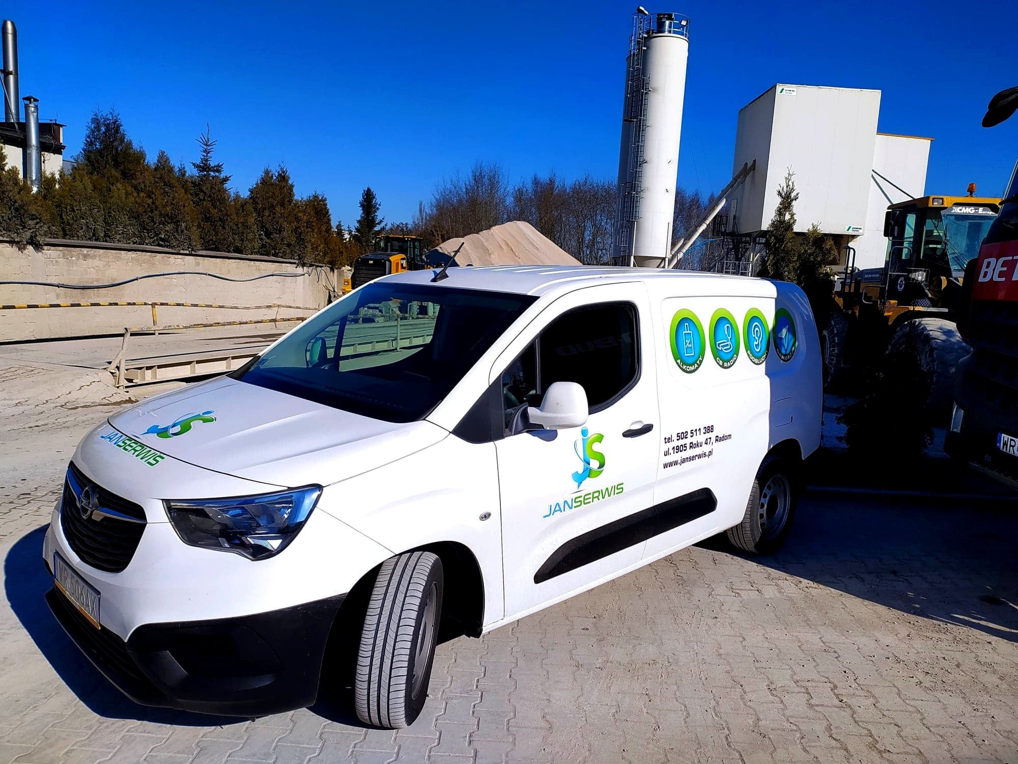 Biała furgonetka serwisowa z logo firmy Janserwis na tle zakładu przemysłowego z silosami i kopcami piasku, widoczny również ciężki sprzęt budowlany.