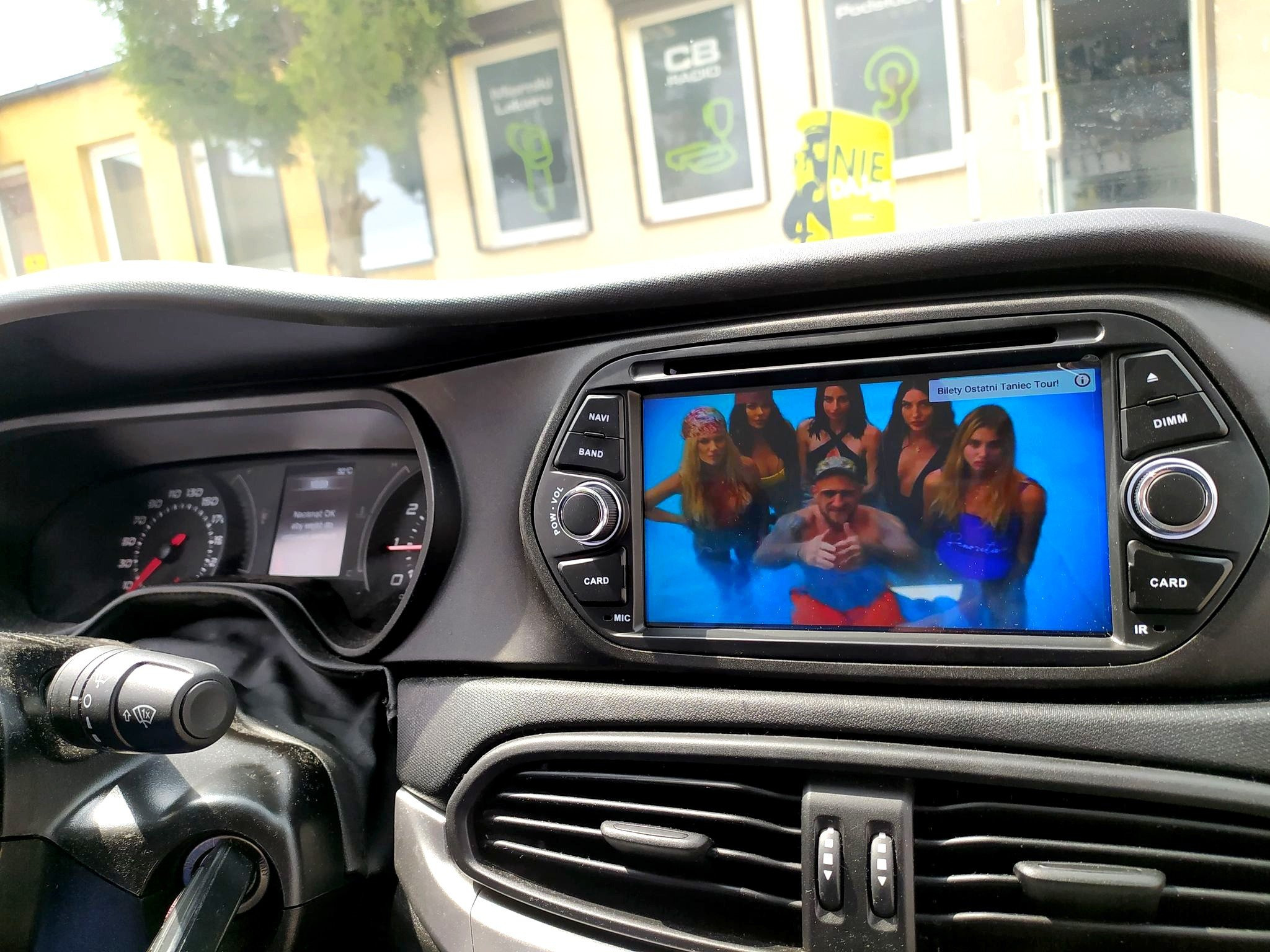 Wnętrze samochodu z włączonym ekranem radia samochodowego, na którym wyświetlany jest teledysk z pływającymi osobami.
