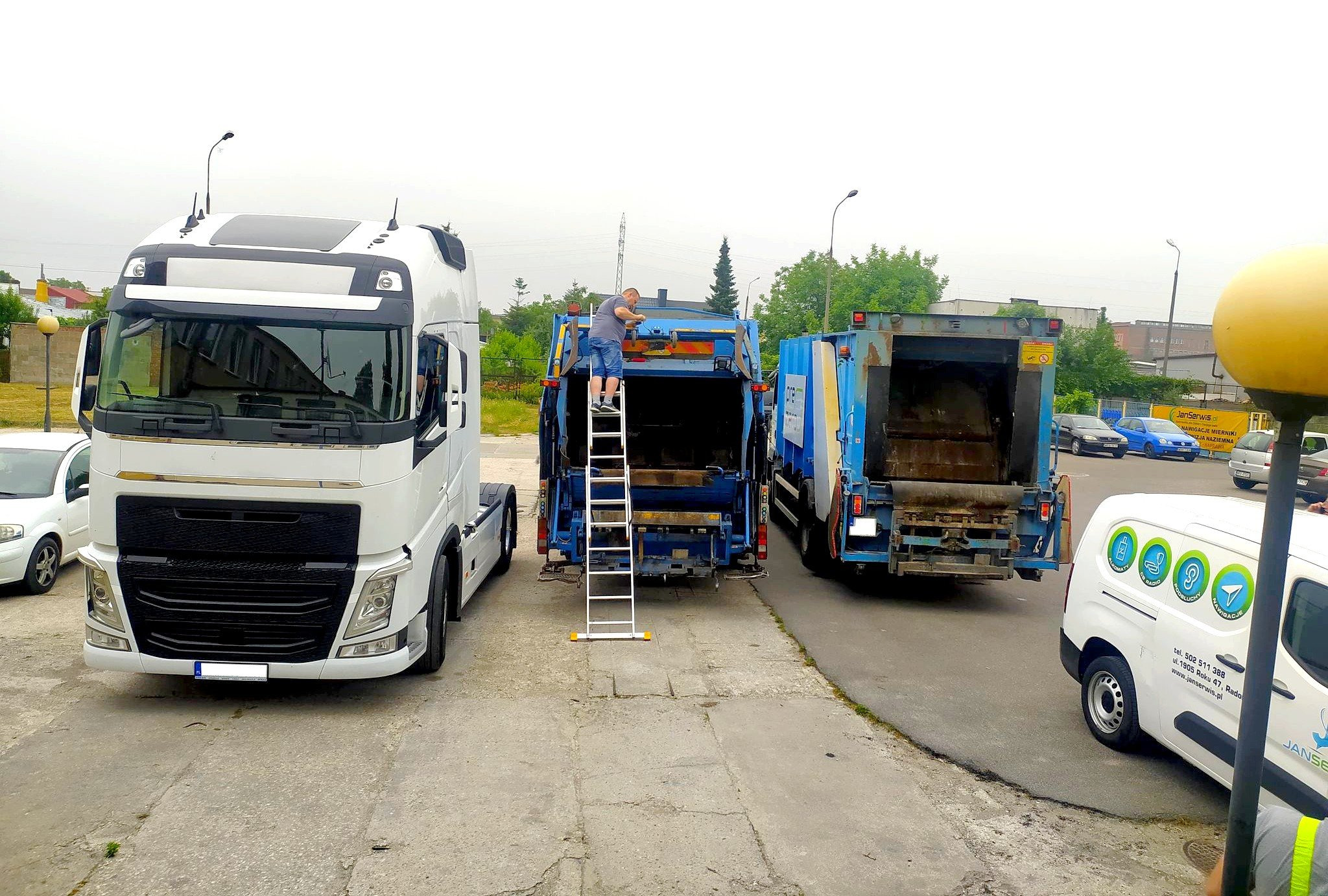 Serwisant na drabinie przy dwóch otwartych śmieciarkach, biała ciężarówka z przodu, firmowy van z reklamą serwisu obok, szare niebo.
