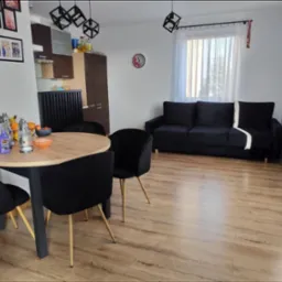 Jasne wnętrze pokoju dziennego z drewnianą podłogą, okrągłym stołem z czarnymi krzesłami, czarną sofą pod oknem z białymi zasłonami i wiszącymi lampami w kształcie kostek