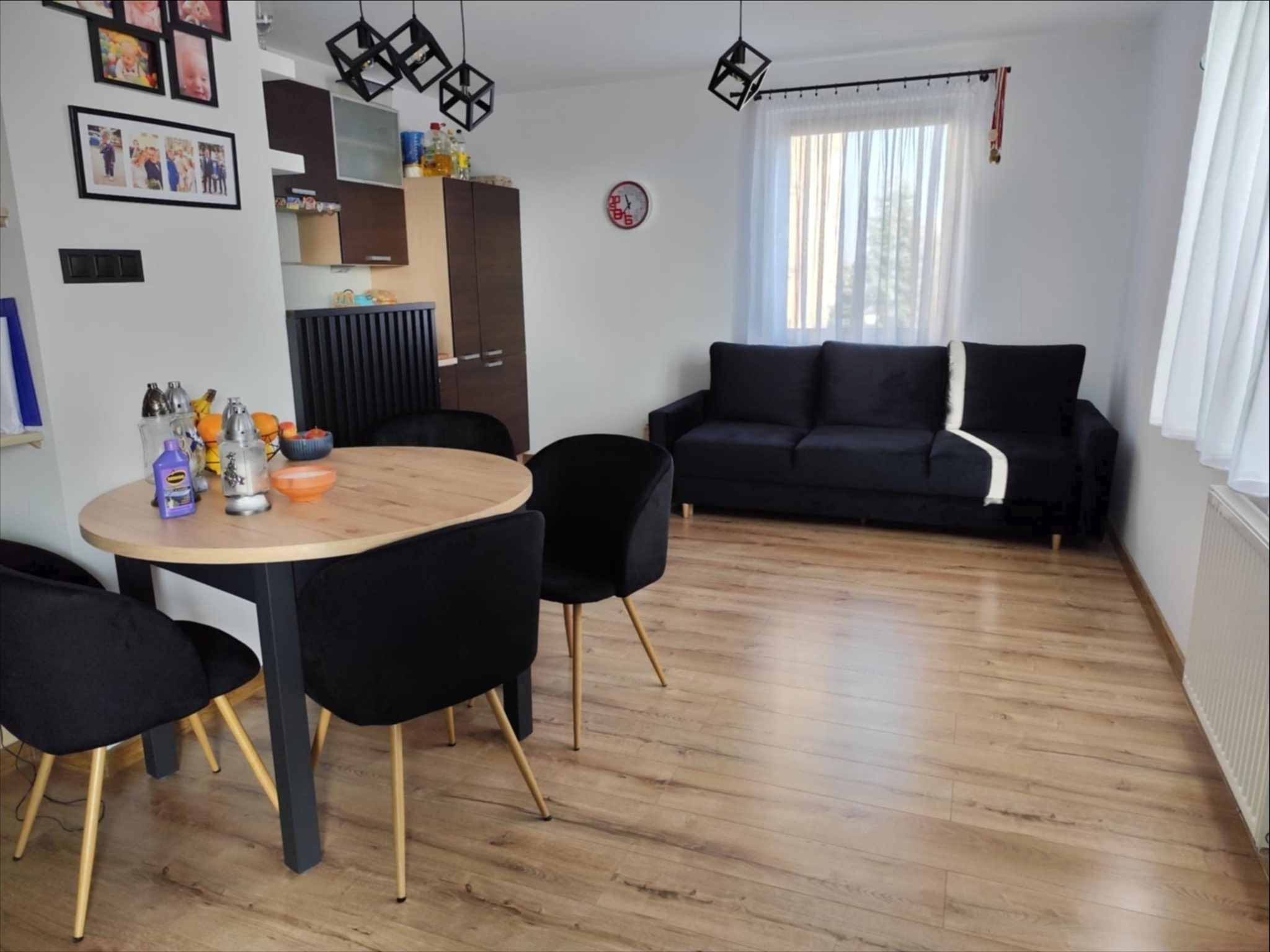 Jasne wnętrze pokoju dziennego z drewnianą podłogą, okrągłym stołem z czarnymi krzesłami, czarną sofą pod oknem z białymi zasłonami i wiszącymi lampami w kształcie kostek