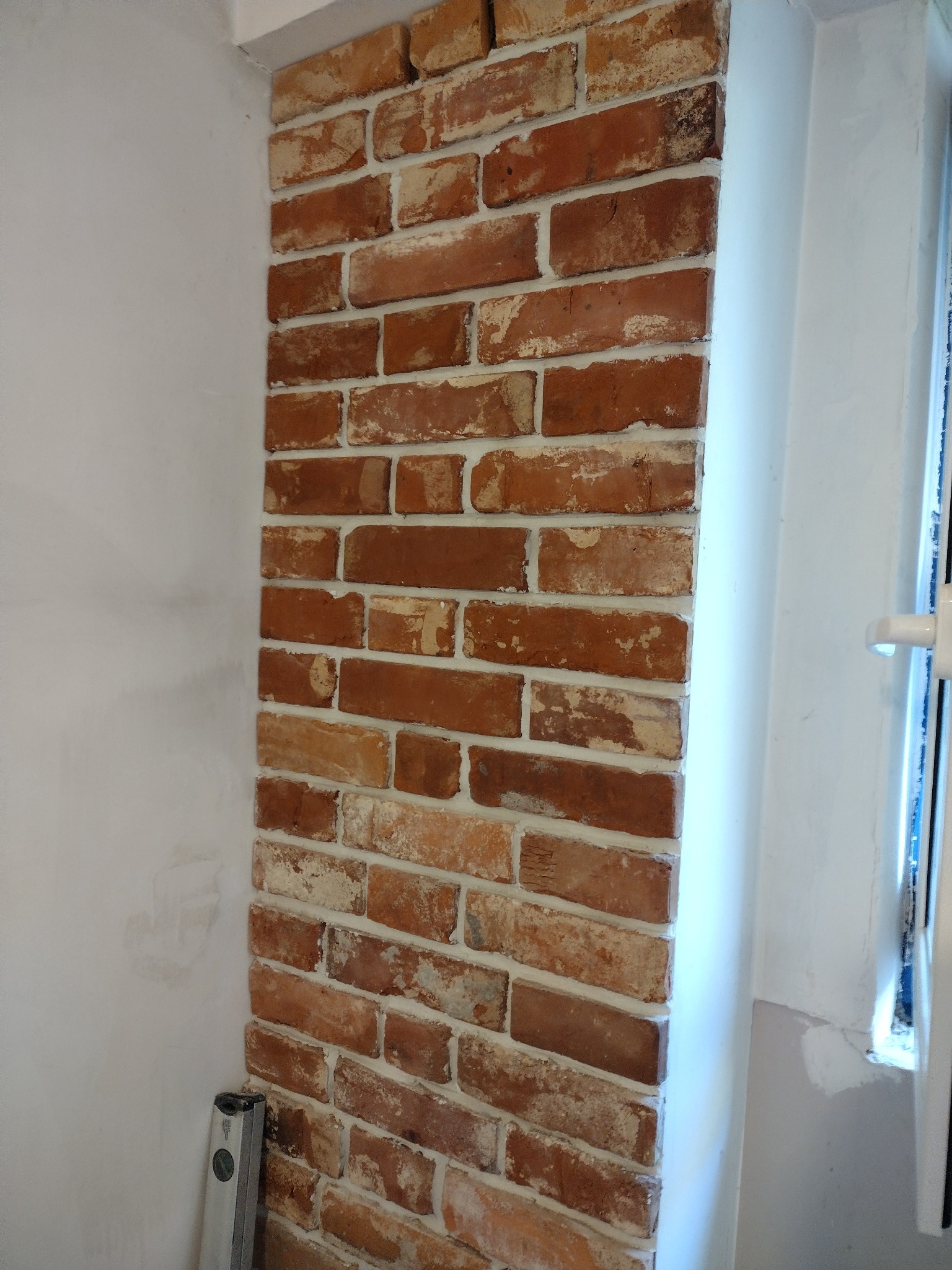 Fragment ściany z dekoracyjnej cegły wewnątrz pomieszczenia, widoczny poziomica przy podłodze, białe wykończenie przy oknie.