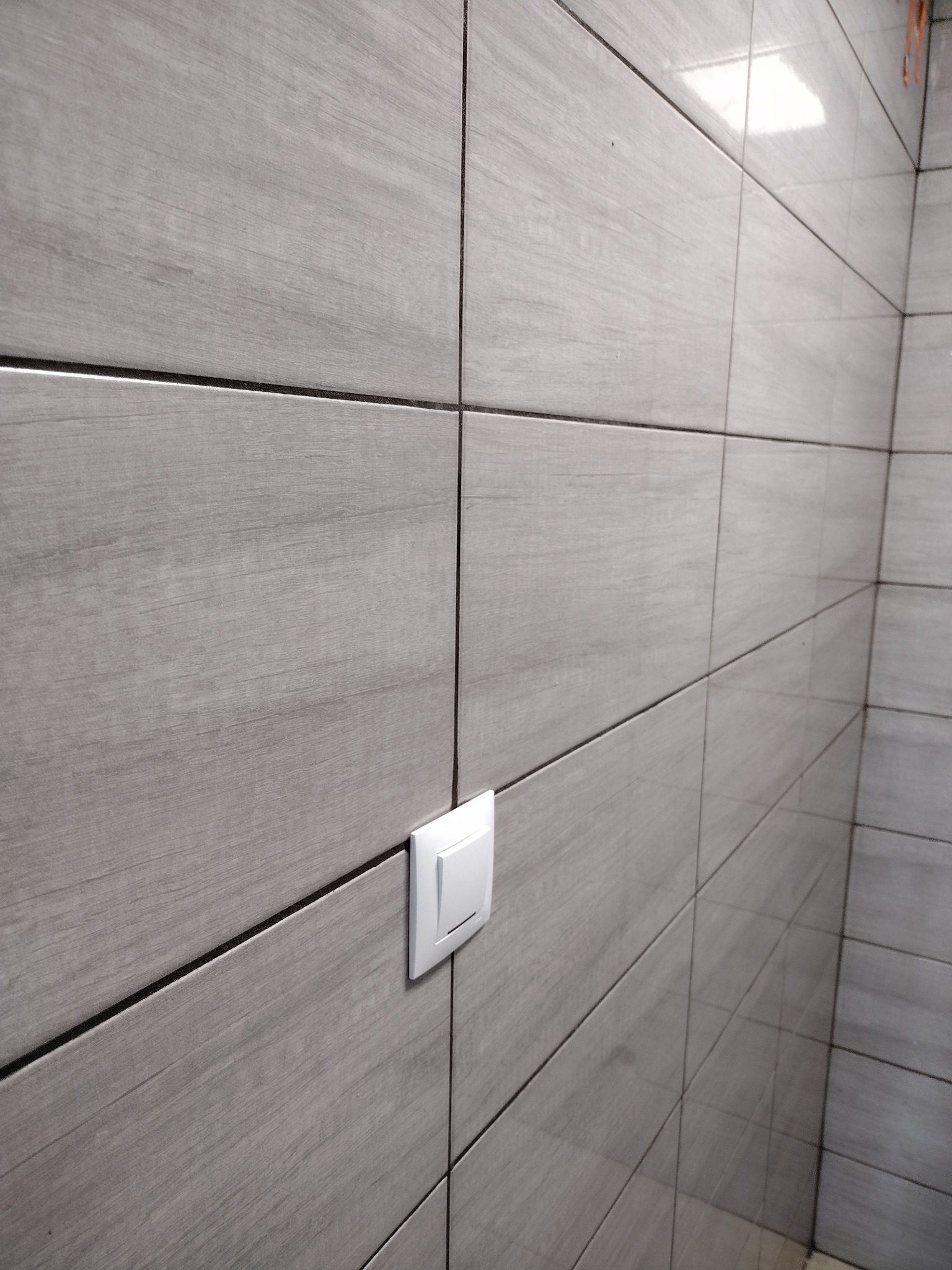 Ściana wyłożona szarymi, podłużnymi płytkami z fakturą drewna i włącznikiem światła.