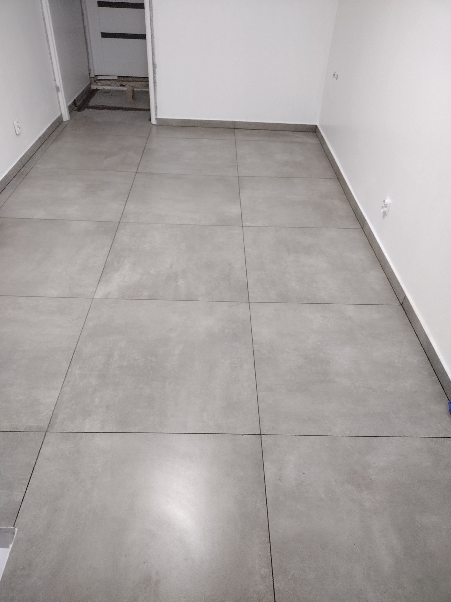 Szare, matowe płytki podłogowe ułożone w nowym pomieszczeniu z białymi ścianami i widocznym wejściem do innego pomieszczenia w tle.