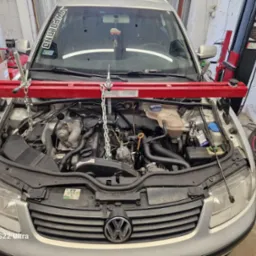 Srebrny samochód z otwartą maską w warsztacie, silnik podtrzymywany przez czerwony poprzeczny wspornik z łańcuchami, widoczne koło z różową felgą i elementy zawieszenia obok.