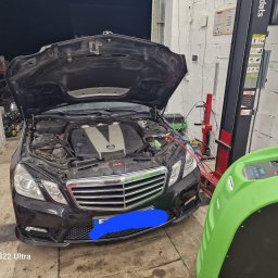 Mechanika Pojazdowa Grzegorz Pater - Czarny Mercedes z otwartą maską w warsztacie, podłączony do zielonego urządzenia do klimatyzacji, widoczny podnośnik samochodowy i narzędzia.