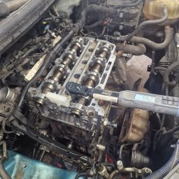 Mechanika Pojazdowa Grzegorz Pater - Silnik samochodowy z otwartą głowicą, widoczne wałki rozrządu, klucz dynamometryczny Snap-on ustawiony na 59.7 Nm, przewody i elementy osprzętu silnika, w tle dokumentacja techniczna.