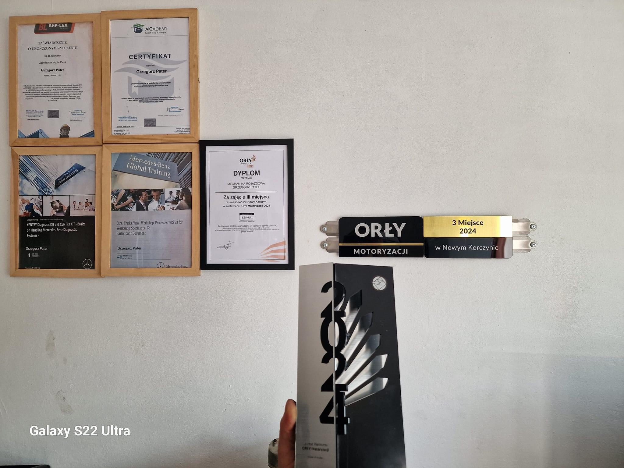 Ściana z dyplomami i certyfikatami warsztatu samochodowego, w tym nagroda Orły Motoryzacji 2024 za zajęcie 3 miejsca, świadectwa ukończonych szkoleń z zakresu diagnostyki i klimatyzacji...