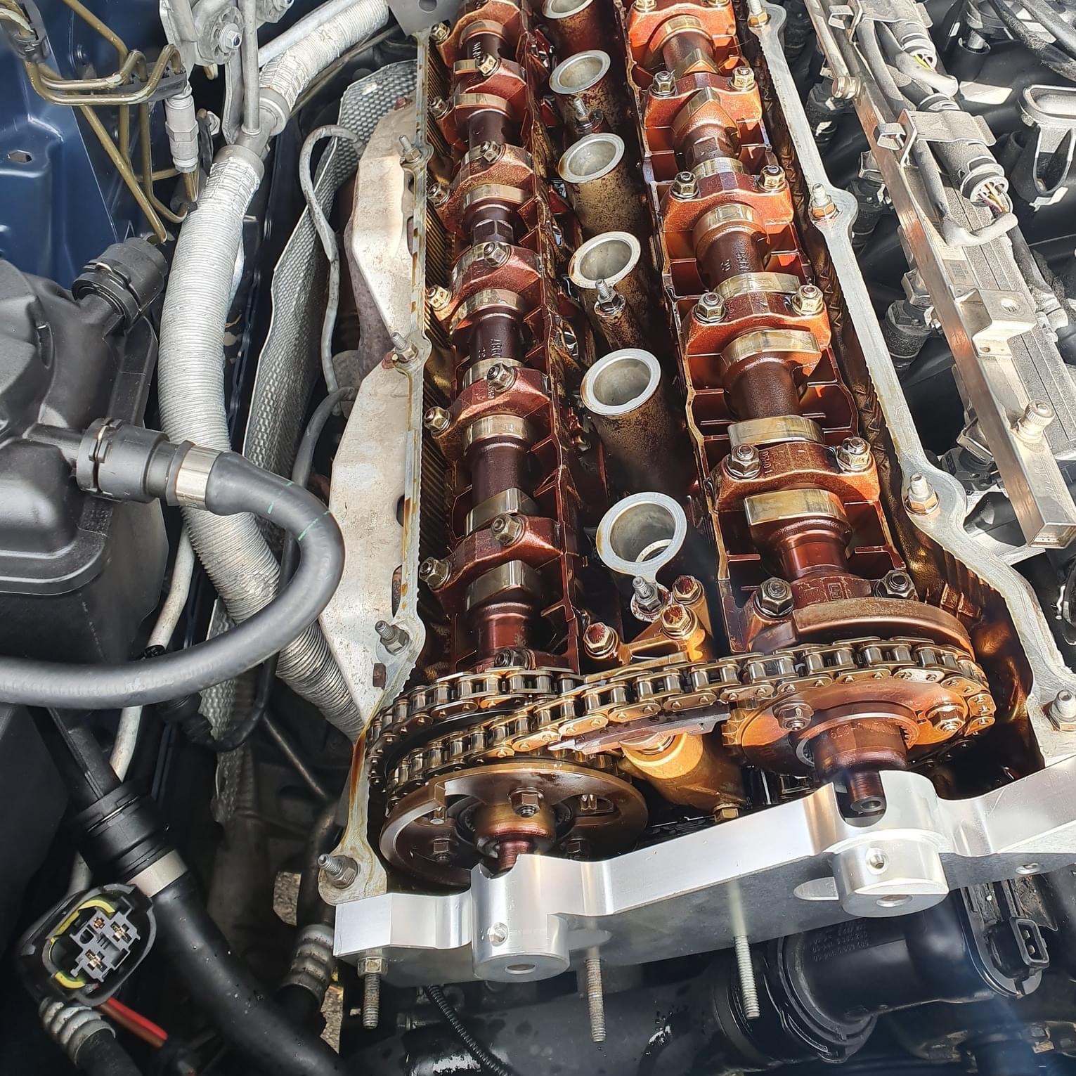 Otwarty silnik samochodowy z widocznymi wałkami rozrządu, łańcuchem rozrządu i elementami mocującymi, ukazujący wewnętrzne mechanizmy w trakcie naprawy lub diagnostyki.