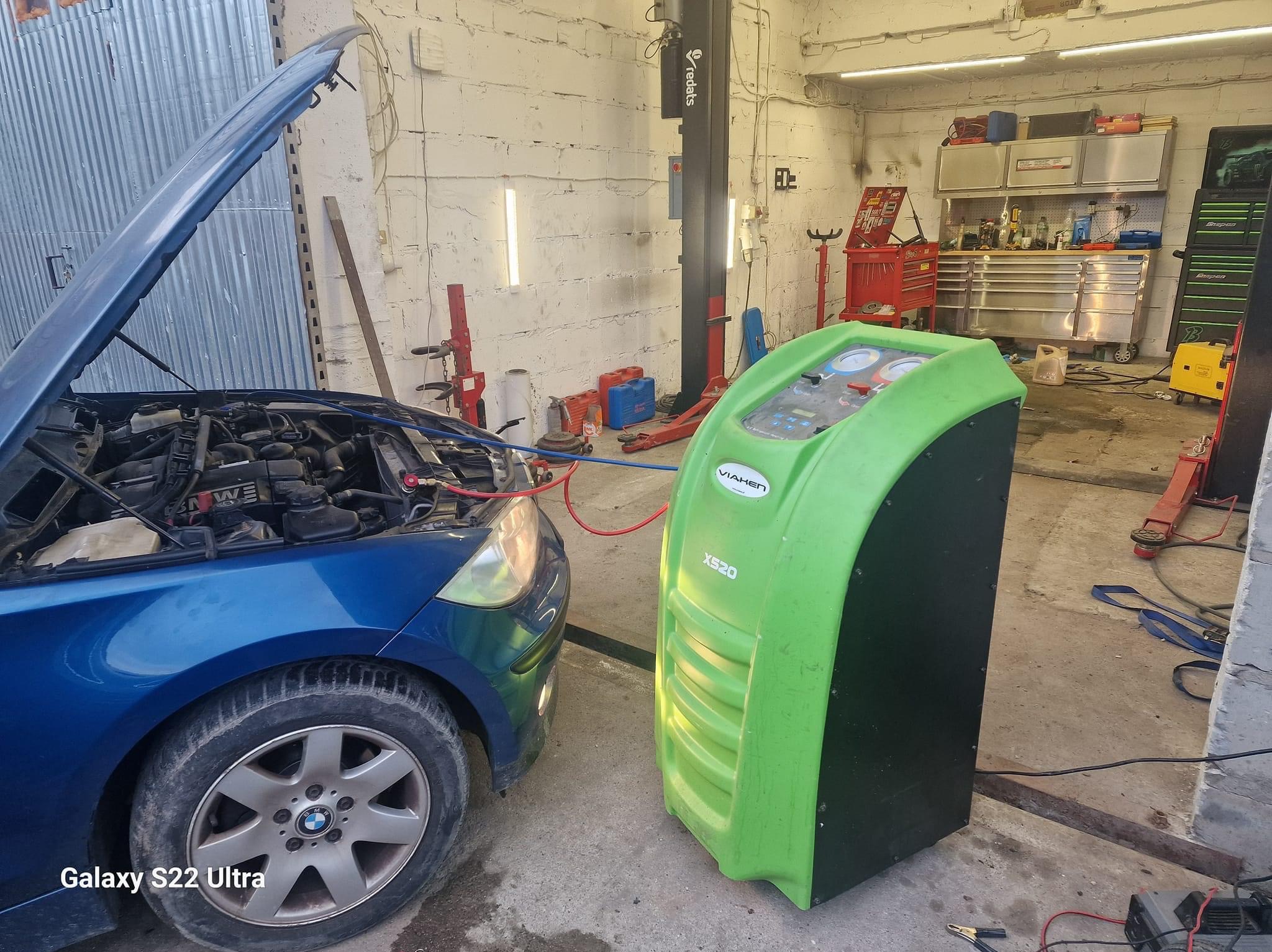 Niebieski samochód z otwartą maską podłączony do zielonego urządzenia do serwisowania klimatyzacji w warsztacie samochodowym.