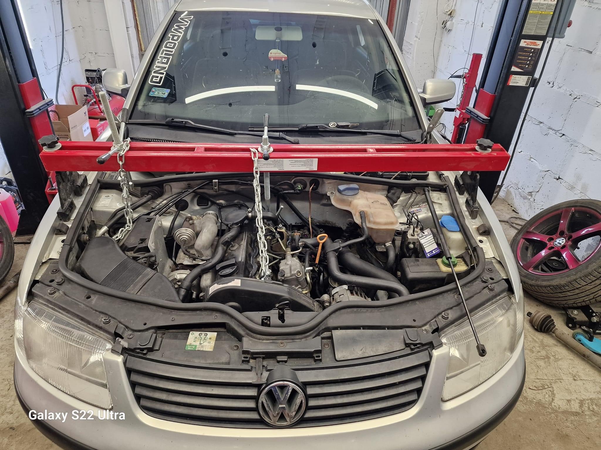 Srebrny samochód z otwartą maską w warsztacie, silnik podtrzymywany przez czerwony poprzeczny wspornik z łańcuchami, widoczne koło z różową felgą i elementy zawieszenia obok.