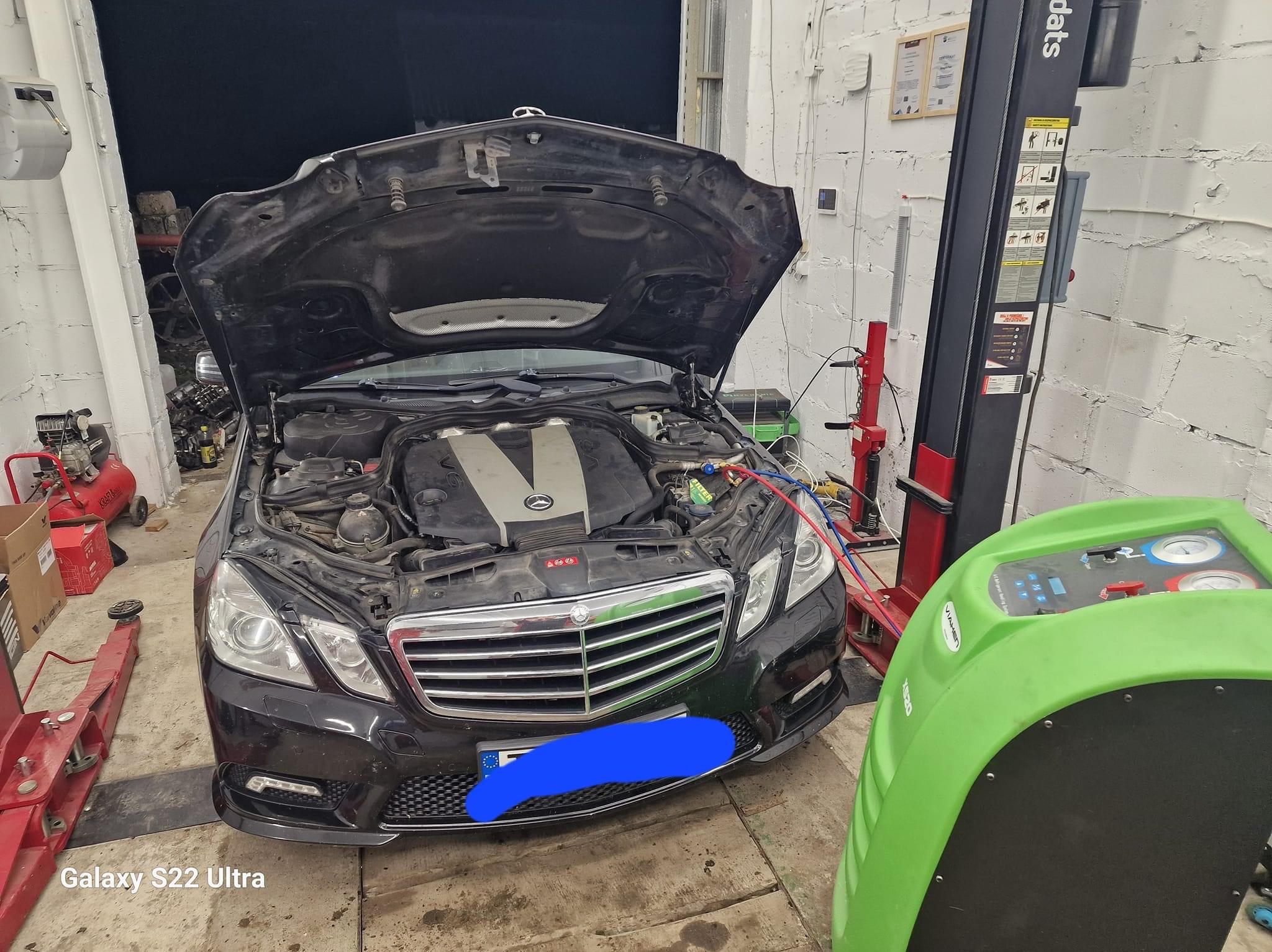 Czarny Mercedes z otwartą maską w warsztacie, podłączony do zielonego urządzenia do klimatyzacji, widoczny podnośnik samochodowy i narzędzia.