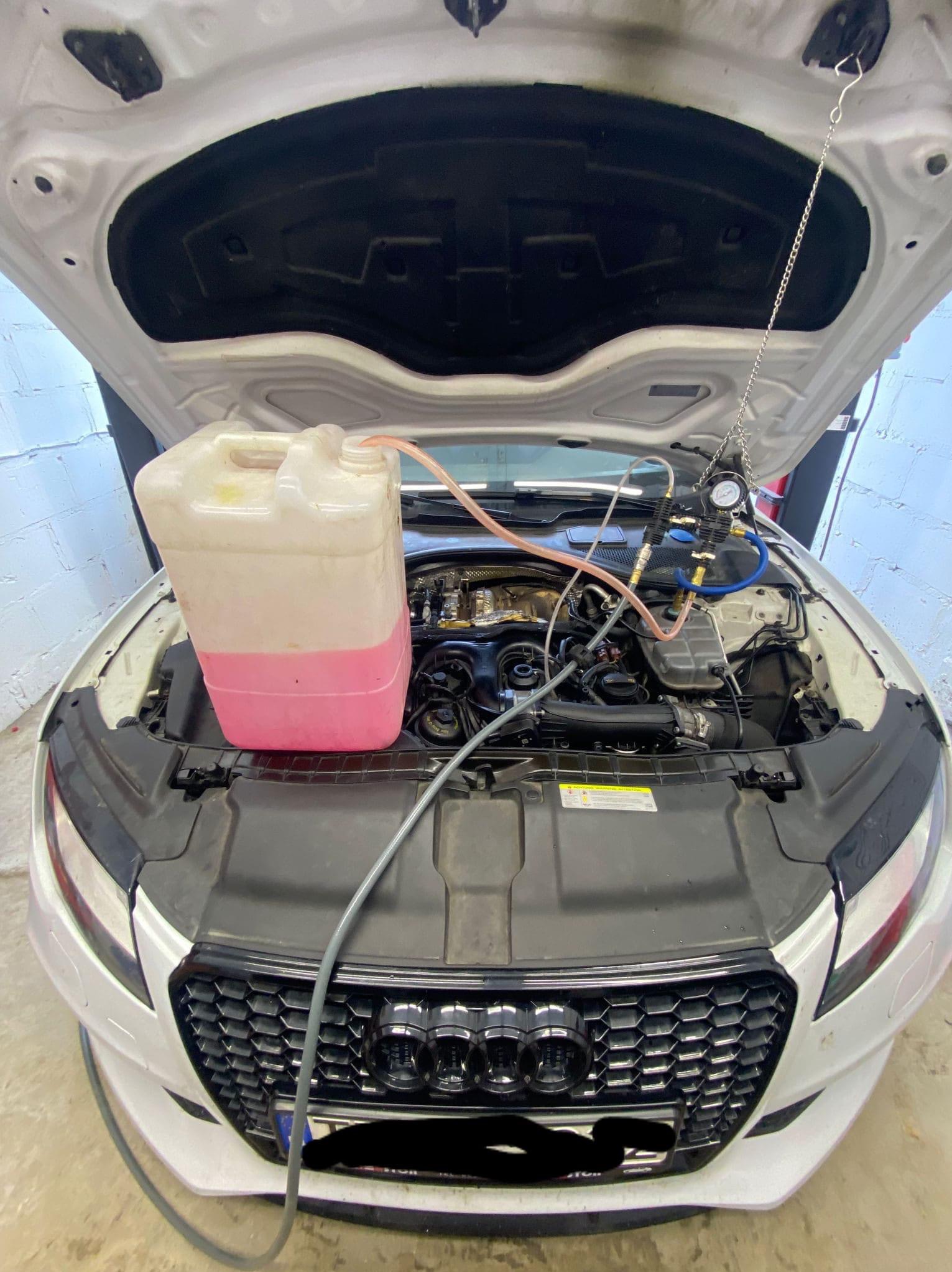 Białe Audi z otwartą maską podczas wymiany płynu chłodniczego; widoczny pojemnik z różowym płynem i podłączone przewody.