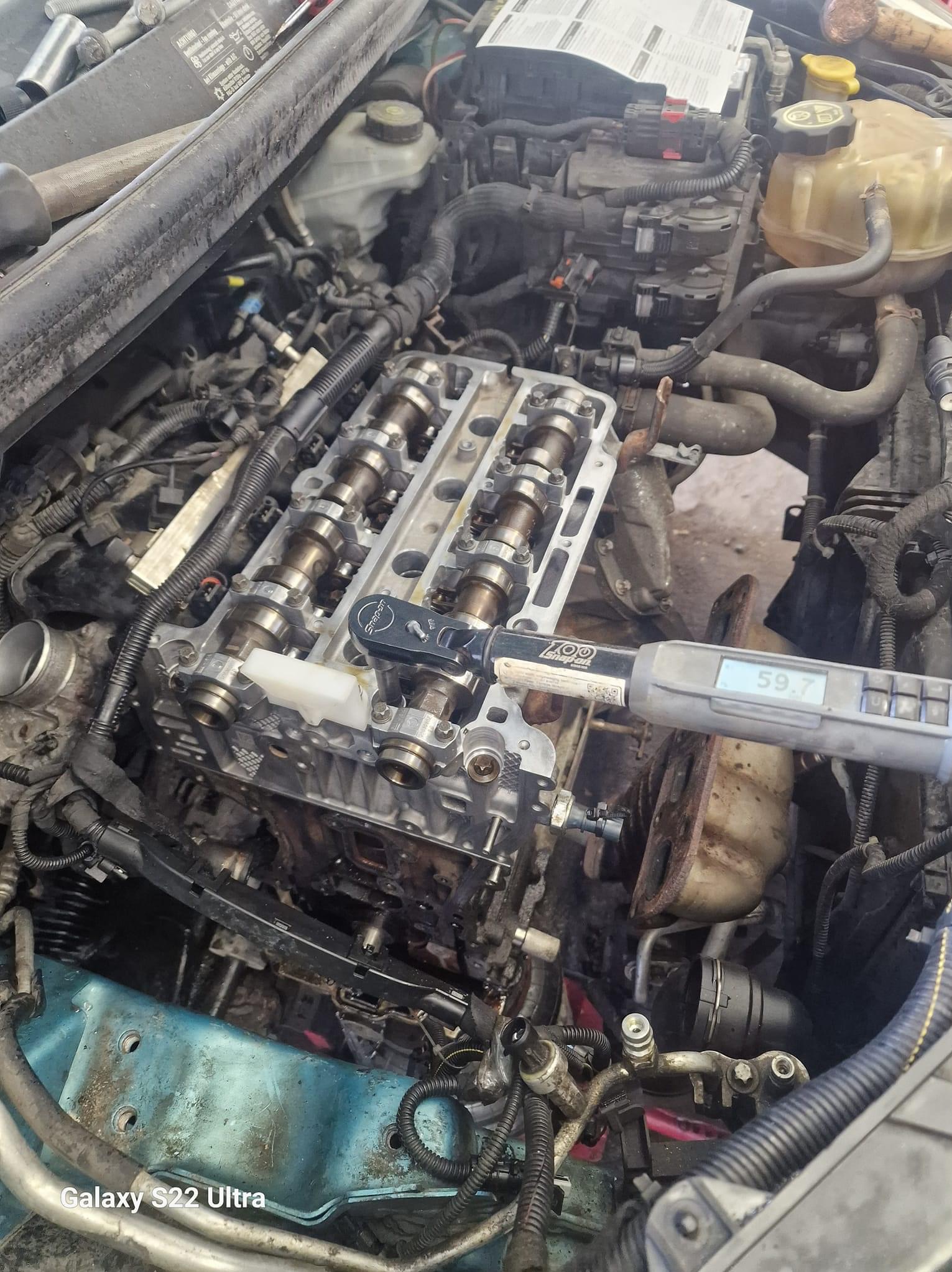 Silnik samochodowy z otwartą głowicą, widoczne wałki rozrządu, klucz dynamometryczny Snap-on ustawiony na 59.7 Nm, przewody i elementy osprzętu silnika, w tle dokumentacja techniczna.