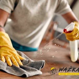 WAŻKA Usługi Sprzątające - Osoba w żółtych rękawiczkach spryskuje i wyciera drewniany blat szarą ściereczką, widoczne drobne krople rozpylonego płynu.