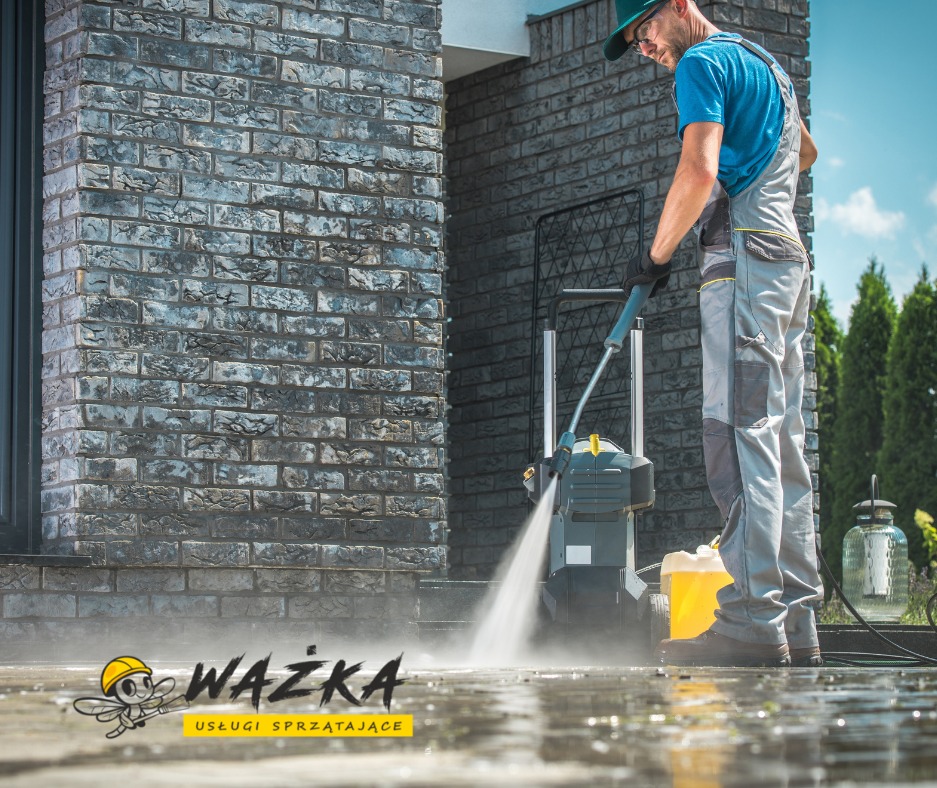 Pracownik w kombinezonie i czapce myje ciśnieniowo chodnik przed budynkiem z ciemnej cegły, widoczny żółty pojemnik z płynem i urządzenie czyszczące.