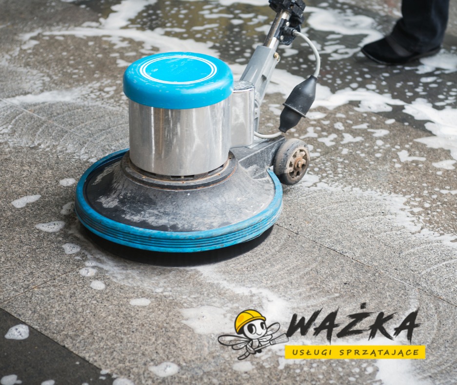 Automat szorujący z niebieskimi akcentami czyszczący szarą posadzkę, widoczne ślady piany i logo firmy sprzątającej w prawym dolnym rogu.