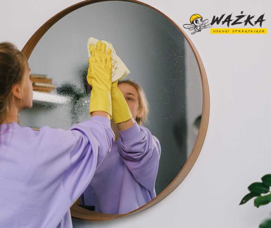 Okrągłe lustro z drewnianą ramą czyszczone żółtą szmatką przez osobę w fioletowej bluzie i żółtych rękawicach ochronnych; widoczne ślady wilgoci na powierzchni lustra.