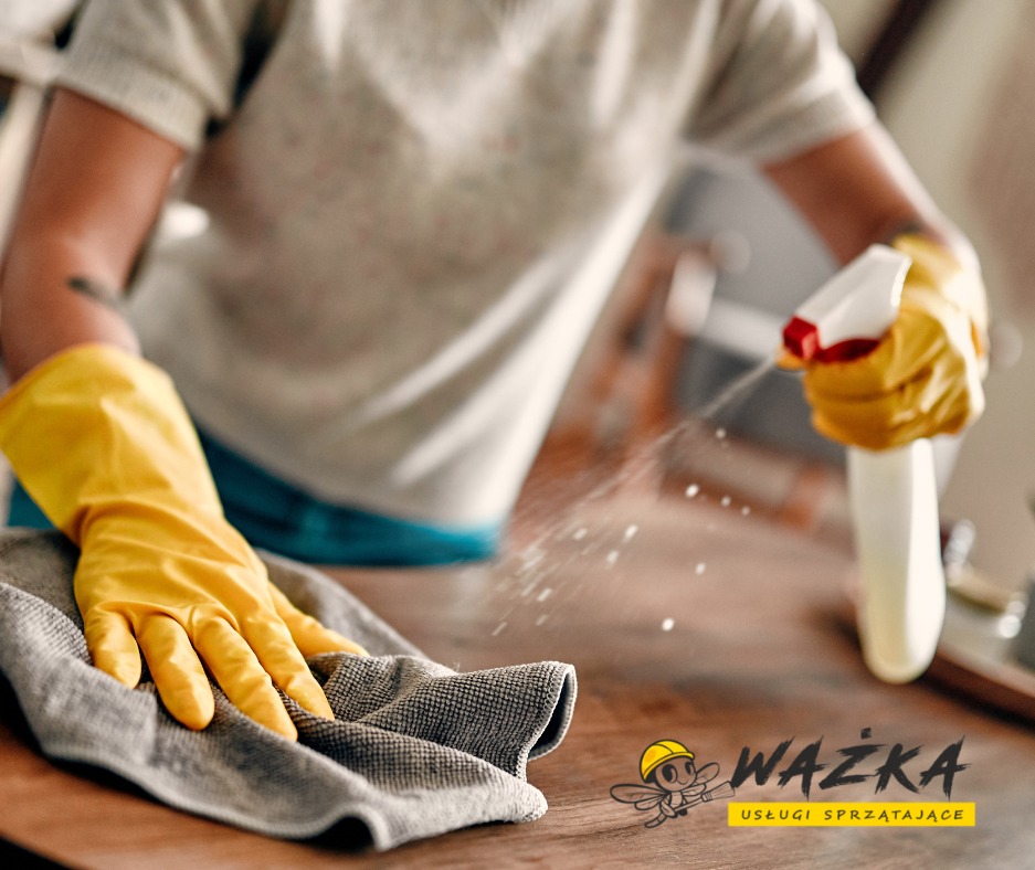 Osoba w żółtych rękawiczkach spryskuje i wyciera drewniany blat szarą ściereczką, widoczne drobne krople rozpylonego płynu.