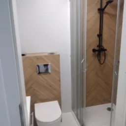 Nowoczesna łazienka z kabiną prysznicową i podwieszaną toaletą, wykończona płytkami w jodełkę. Minimalistyczny design, czarna armatura.