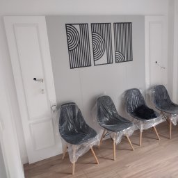 Przemysław Stępiń - Białe drzwi z ościeżnicą, ozdobne panele ścienne i cztery krzesła z tworzywa sztucznego zabezpieczone folią ochronną w jasnym, odnowionym wnętrzu.