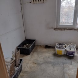 Przemysław Stępiń - Pokój w trakcie renowacji: wiaderka z resztkami płytek, gruz w pojemniku, grzejnik na ścianie, okno z widokiem na drzewa. Przygotowania do prac.
