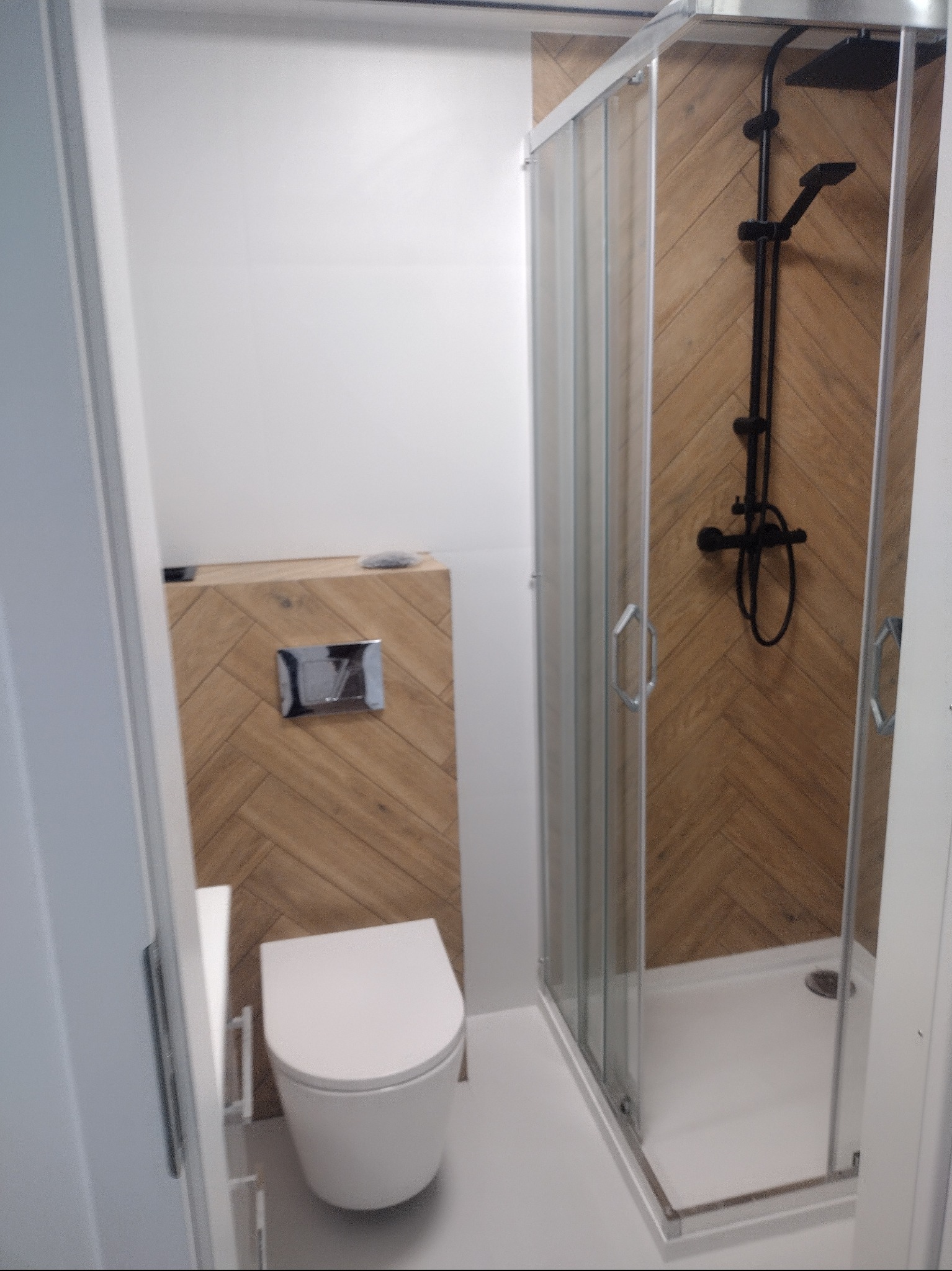 Nowoczesna łazienka z kabiną prysznicową i podwieszaną toaletą, wykończona płytkami w jodełkę. Minimalistyczny design, czarna armatura.