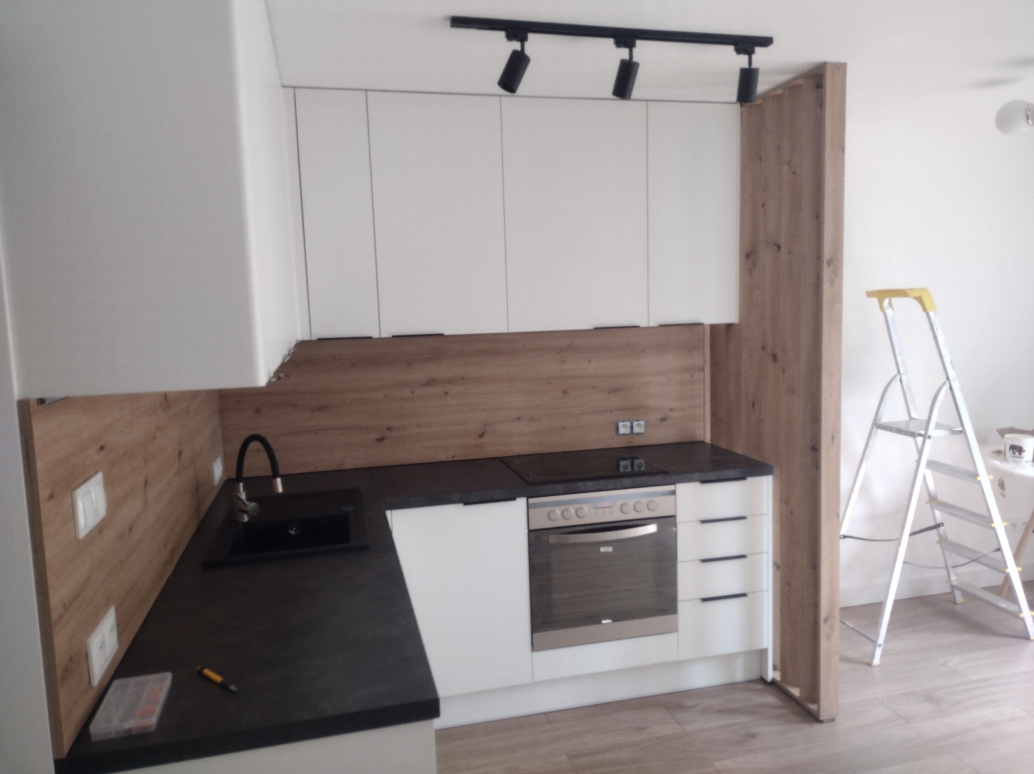 Nowoczesna kuchnia z białymi szafkami, drewnianym panelem i czarnym blatem. W tle drabina, sugerująca prace wykończeniowe. Oświetlenie sufitowe punktowe.