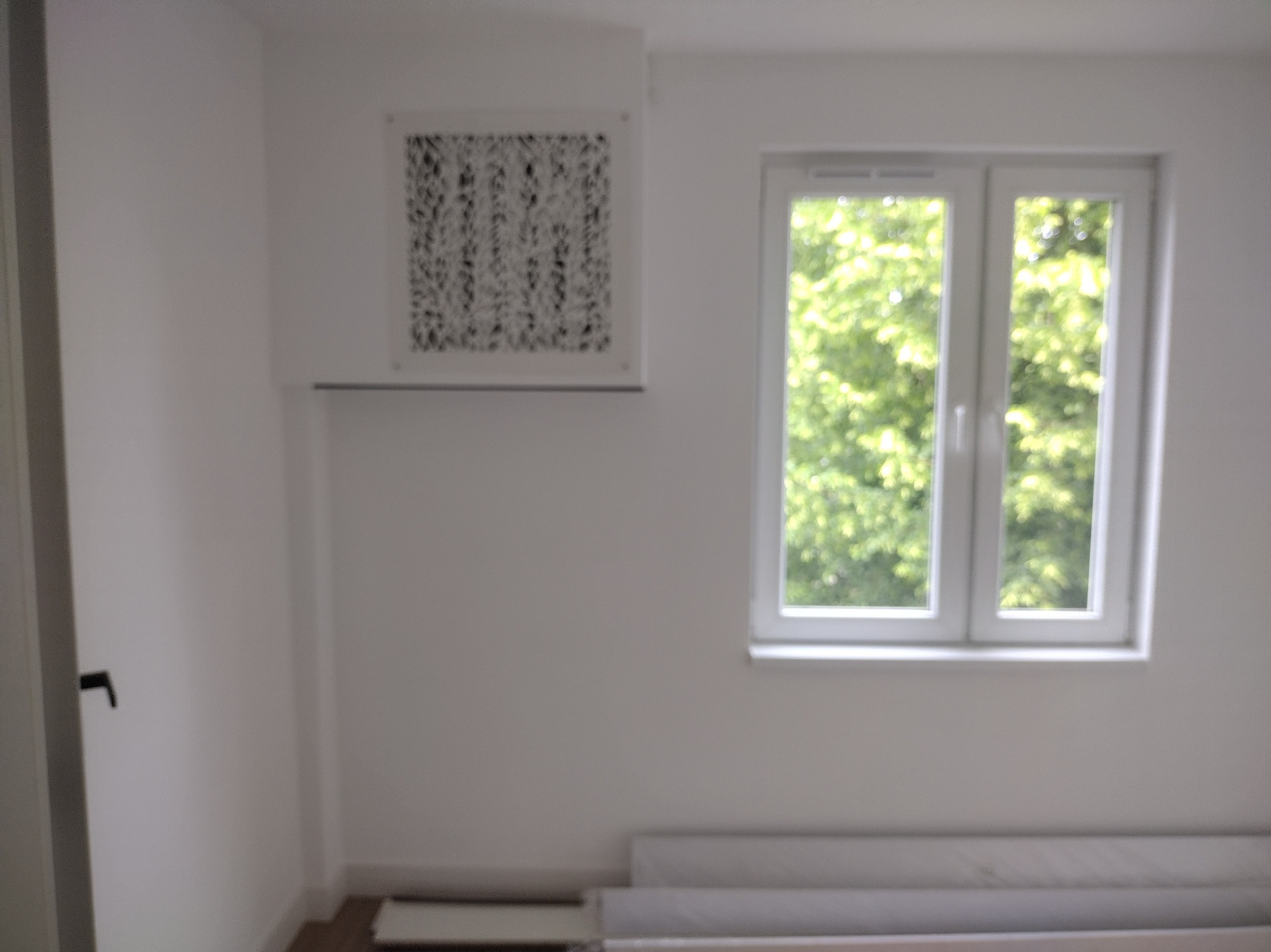 Białe wnętrze pokoju z oknem i dekoracyjnym panelem ściennym. Na podłodze leżą zwinięte dywany. Ściany świeżo pomalowane na biało.