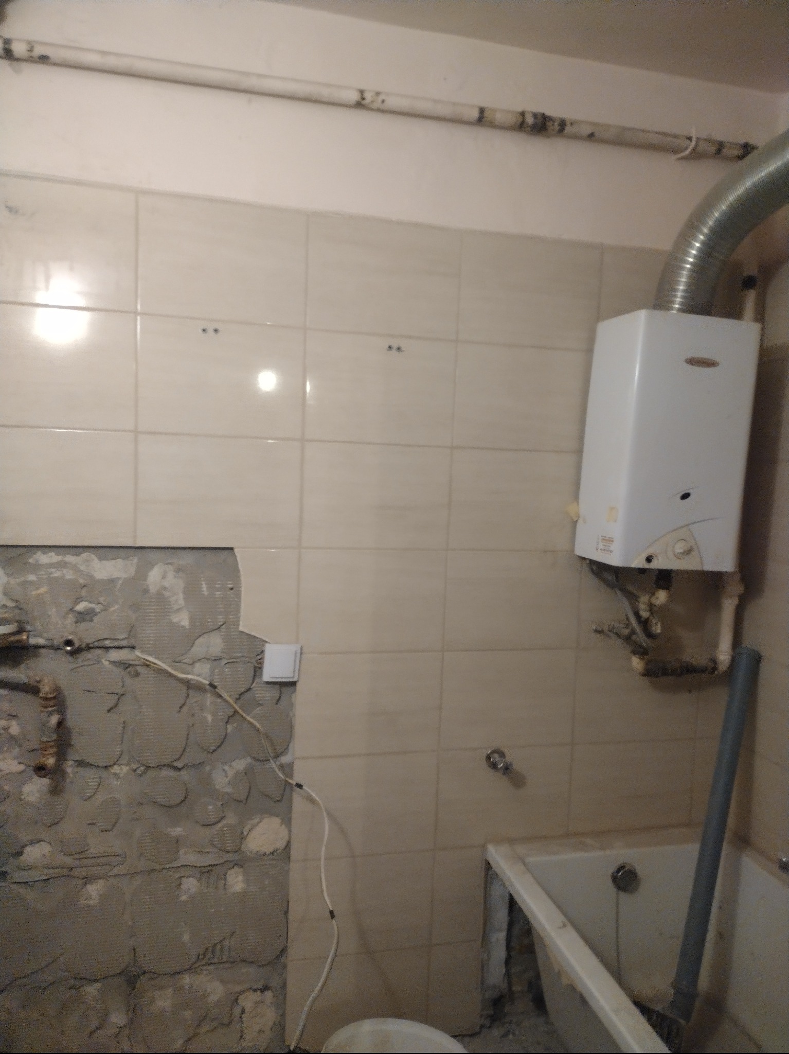 Częściowo zdemontowane płytki w łazience, widoczny fragment ściany z instalacją, bojler, fragment wanny i rury. Etap prac remontowych.