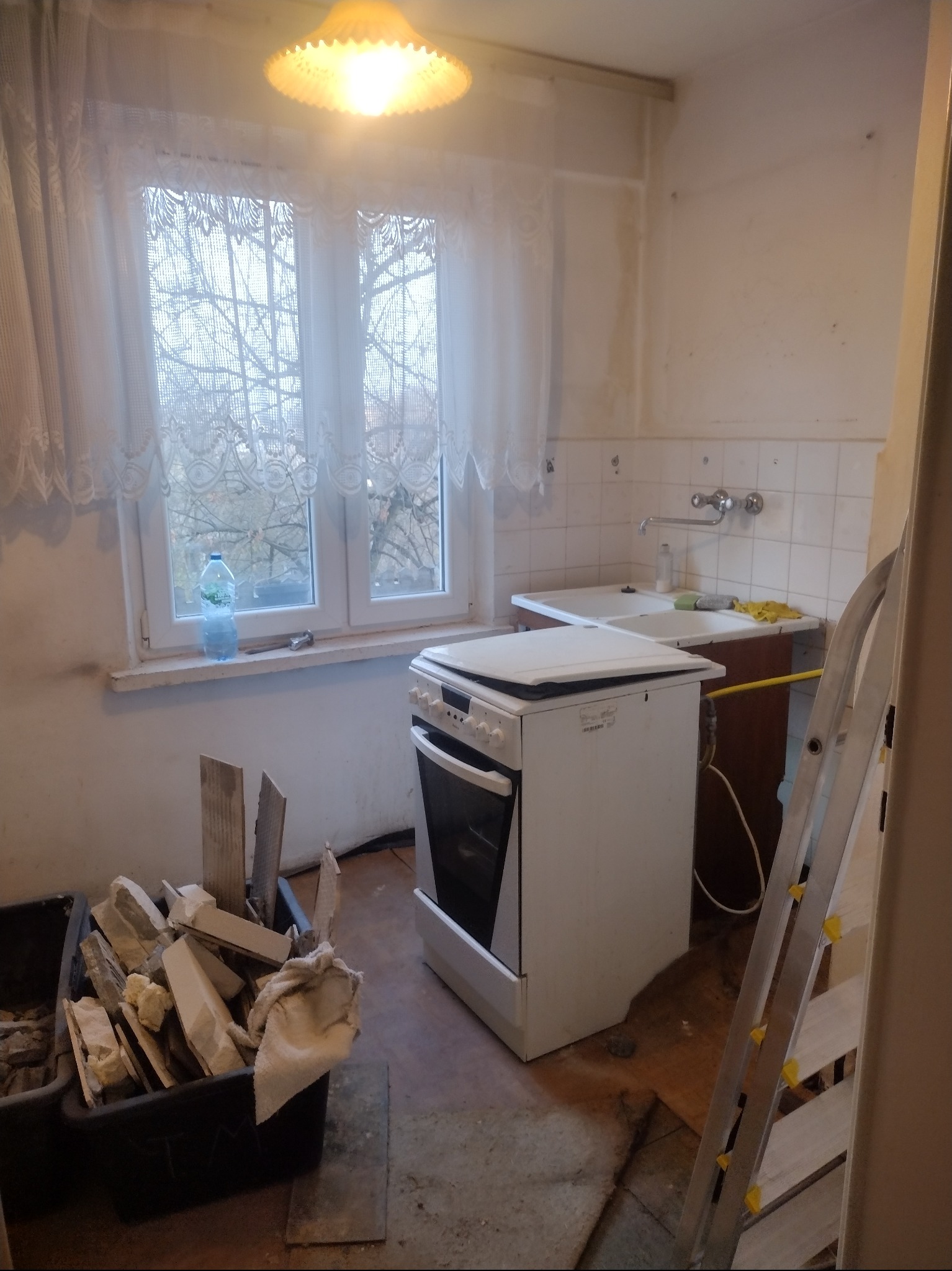 Kuchnia w trakcie demontażu: widoczne stare płytki, okno z firanką, butelka wody, kuchenka i drabina. Przygotowania do remontu.