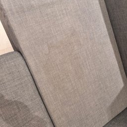 IMA GEFEST SPÓŁKA Z OGRANICZONĄ ODPOWIEDZIALNOŚCIĄ - Fragment sofy z szarej tkaniny o widocznej strukturze, z delikatnym odbarwieniem na siedzisku, ukazujący detale tapicerki i jej fakturę.