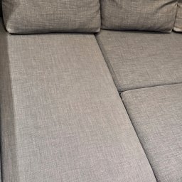 IMA GEFEST SPÓŁKA Z OGRANICZONĄ ODPOWIEDZIALNOŚCIĄ - Szary narożnik z tkaniny o widocznej strukturze, jasne drewniane panele podłogowe w dolnym rogu kadru.