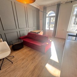 IMA GEFEST SPÓŁKA Z OGRANICZONĄ ODPOWIEDZIALNOŚCIĄ - Nowoczesne wnętrze mieszkania z czerwoną sofą, drewnianą podłogą i szarą ścianą z dekoracyjnymi panelami, oświetlone ciepłym światłem lampy i wpadającym przez okno promieniem słońca.
