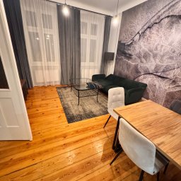 IMA GEFEST SPÓŁKA Z OGRANICZONĄ ODPOWIEDZIALNOŚCIĄ - Ustawny salon z drewnianą podłogą, ciemnozieloną sofą, szklanym stolikiem kawowym i drewnianym stołem z krzesłami na tle ściany z abstrakcyjnym wzorem.