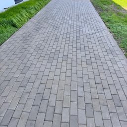 IMA GEFEST SPÓŁKA Z OGRANICZONĄ ODPOWIEDZIALNOŚCIĄ - Ułożona, szara kostka brukowa tworząca ścieżkę wzdłuż budynku z zielonym pasem trawy. Widok z perspektywy.