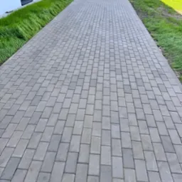 Ułożona, szara kostka brukowa tworząca ścieżkę wzdłuż budynku z zielonym pasem trawy. Widok z perspektywy.