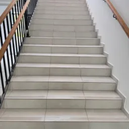 Klatka schodowa z beżowymi płytkami, czarną metalową balustradą i drewnianymi poręczami. Widok z dołu na górę, z widocznym oknem na końcu schodów.