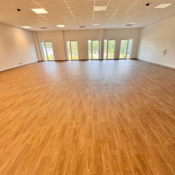 IMA GEFEST SPÓŁKA Z OGRANICZONĄ ODPOWIEDZIALNOŚCIĄ - Jasne, przestronne wnętrze po sprzątaniu. Parkiet z wzorem drewna, białe ściany i sufit z oświetleniem. Widoczne okna i elementy wentylacji.