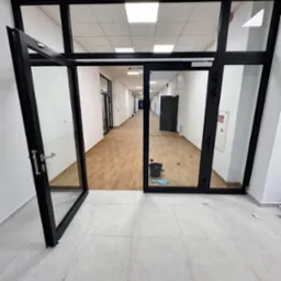 Wnętrze biura po sprzątaniu: otwarte drzwi, widoczny korytarz z czystą podłogą, wiadro i ściereczka w tle, jasne płytki podłogowe. Perspektywa z dołu.