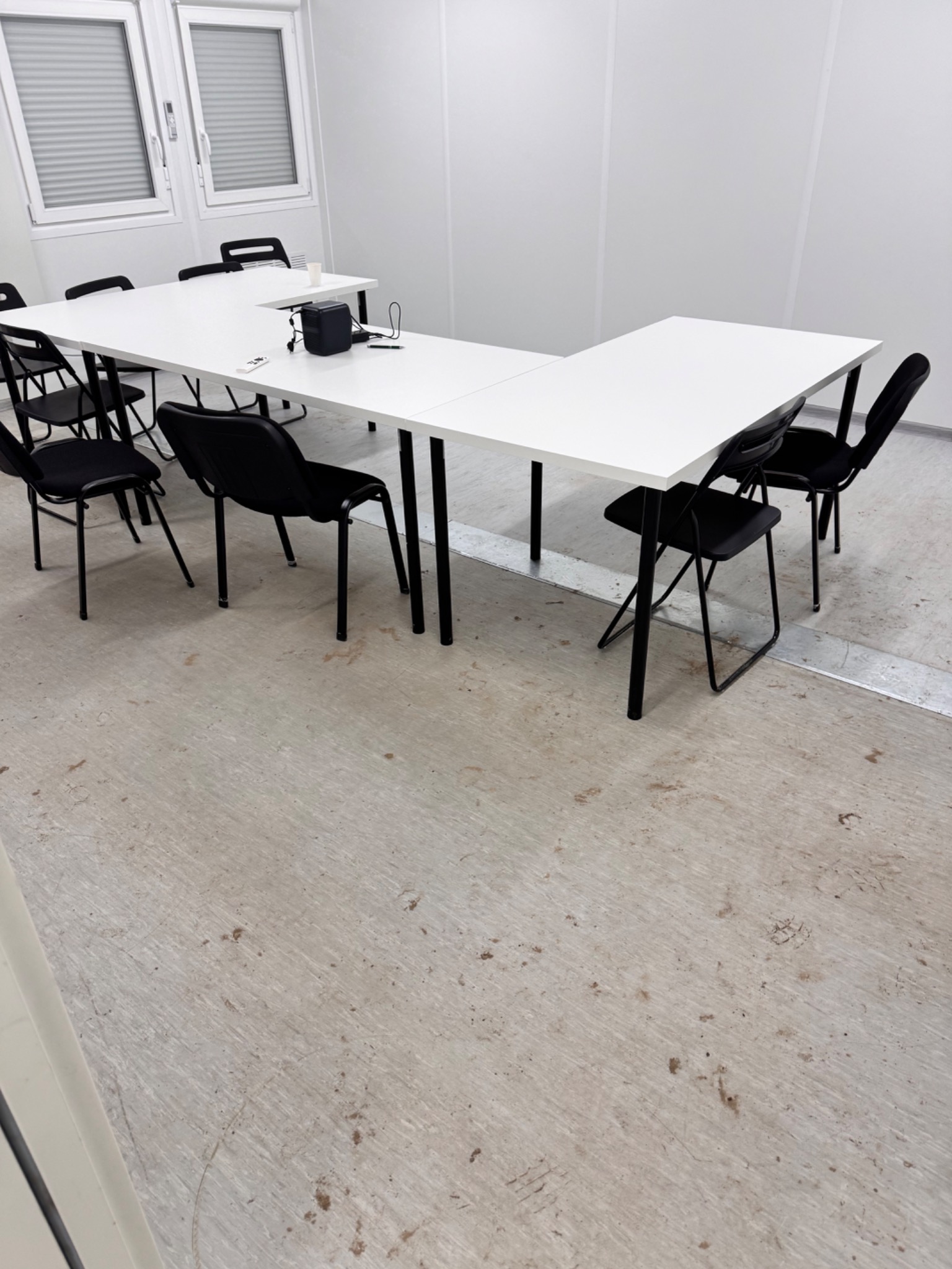 Białe biurko w kształcie litery U z czarnymi krzesłami w jasnym biurze z szarym brudnym linoleum na podłodze.