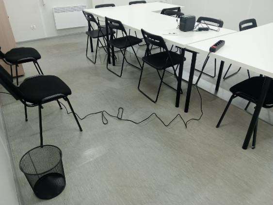 Sala konferencyjna z długim białym stołem, czarnymi krzesłami, grzejnikiem na ścianie, widocznymi kablami na podłodze i koszem na śmieci w kącie.