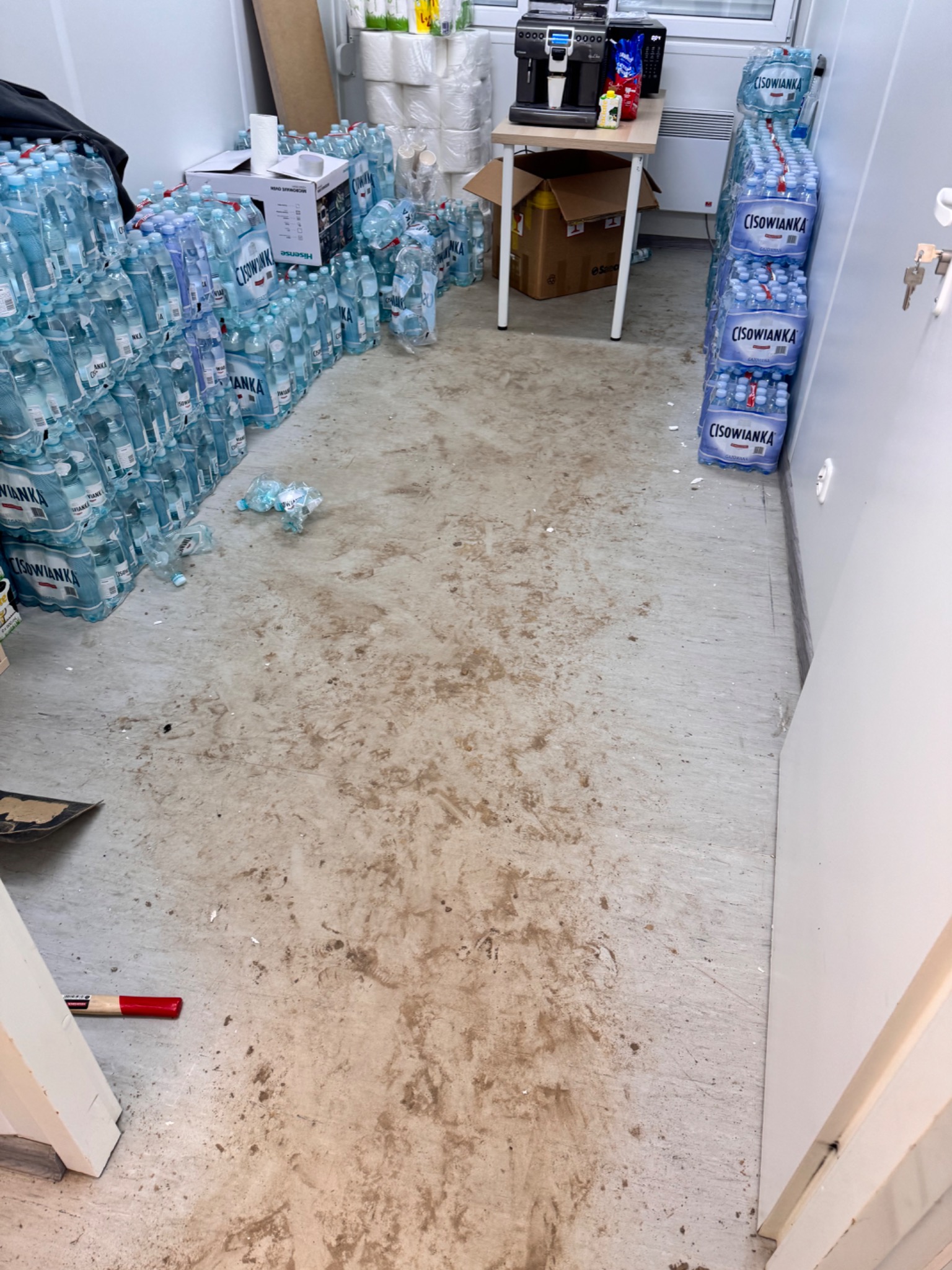 Zabrudzona podłoga w pomieszczeniu magazynowym z zapasem wody mineralnej i papieru toaletowego, widoczne ślady błota i plamy, fragment narzędzia z czerwoną rączką przy progu.