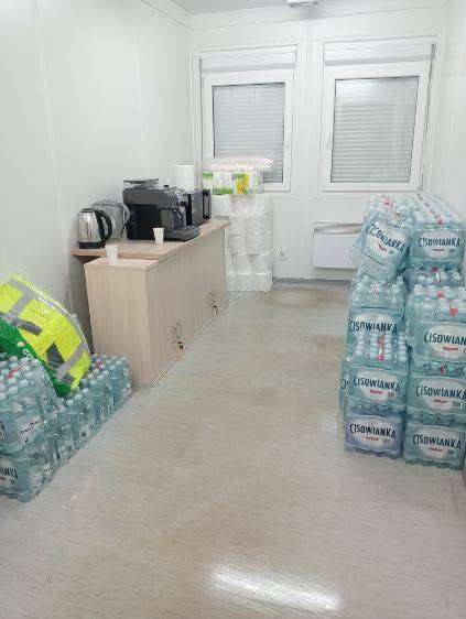 Wnętrze kontenera biurowego z zapasem wody mineralnej Cisowianka, zapasem papieru toaletowego, czajnikiem, ekspresem do kawy i żółtą kamizelką odblaskową na podłodze.
