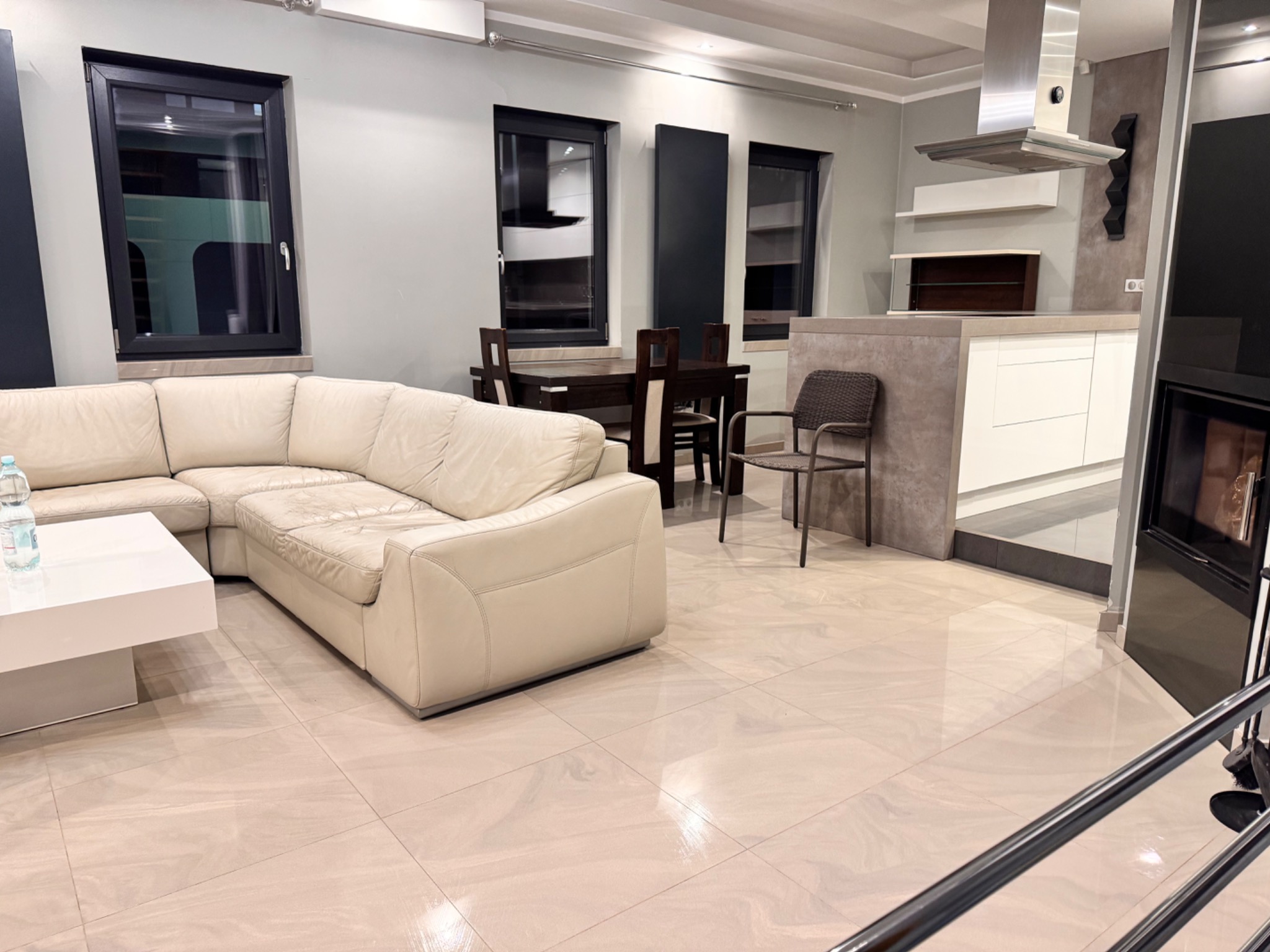 Jasne, przestronne wnętrze salonu z narożną sofą, stołem, jadalnią i aneksem kuchennym w tle, z widoczną podłogą wyłożoną płytkami imitującymi marmur.